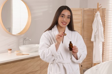 Happy young woman holding bottle and dropper of essential oil in bathroom