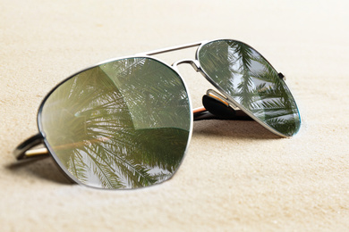 Image of Stylish sunglasses with reflection of palm trees on white sand, closeup 