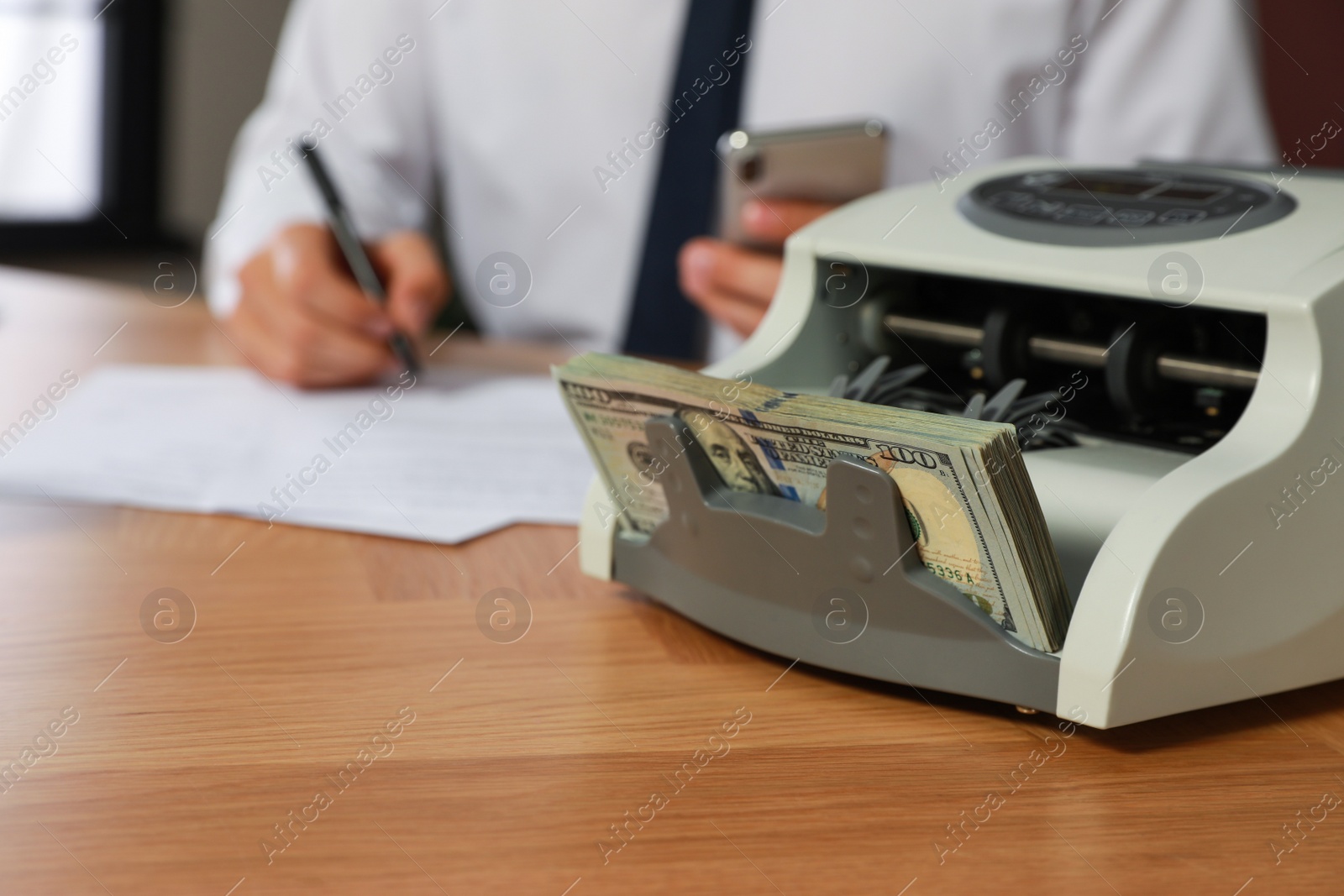 Photo of Modern banknote counter with money and blurred view of man working at wooden table indoors