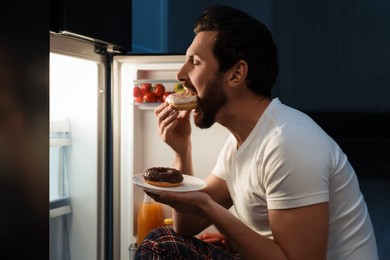 Photo of Man eating donuts near refrigerator in kitchen at night. Bad habit