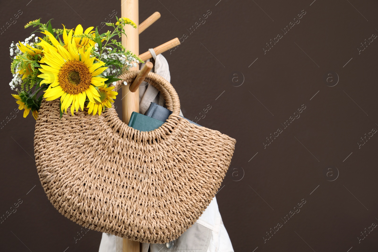 Photo of Stylish beach bag with beautiful bouquet of wildflowers and books hanging on coat stand against brown background. Space for text