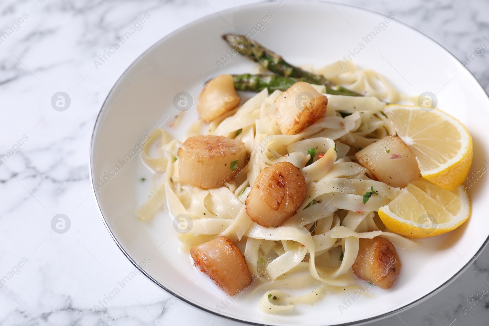 Photo of Delicious scallop pasta with asparagus and lemon on white marble table, closeup