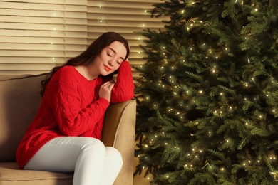 Beautiful woman napping in armchair near Christmas tree with golden lights indoors