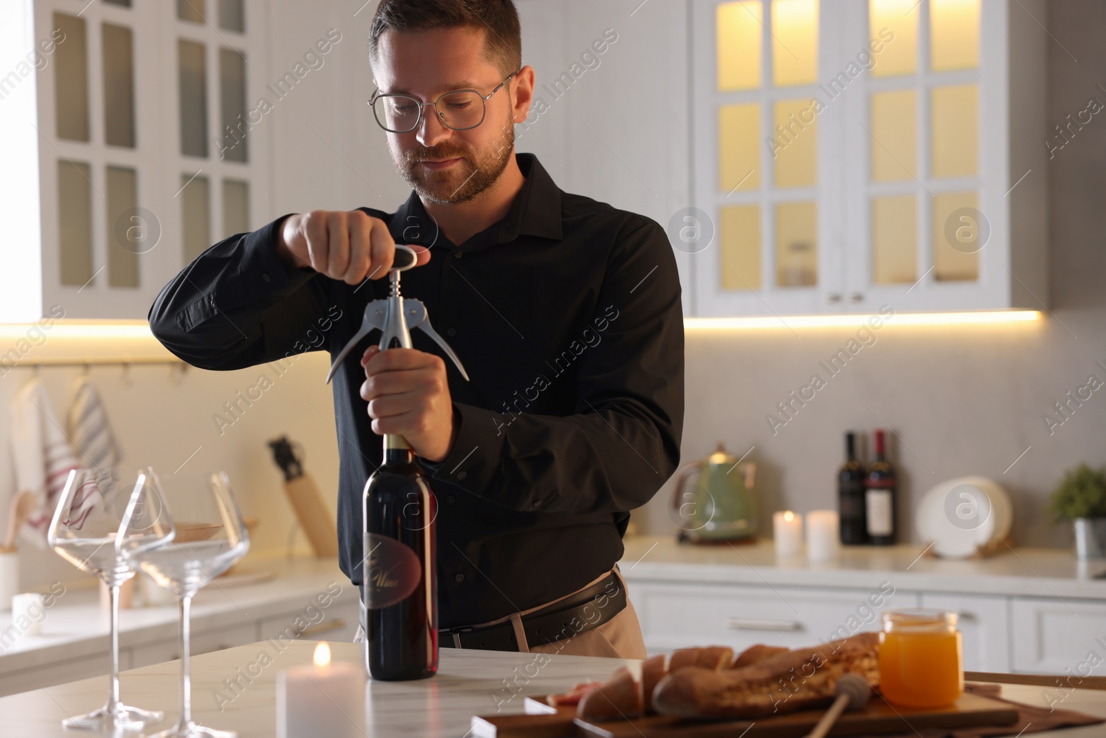 Photo of Romantic dinner. Man opening wine bottle with corkscrew at table in kitchen