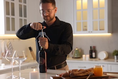 Photo of Romantic dinner. Man opening wine bottle with corkscrew at table in kitchen