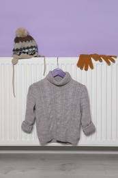 Photo of Heating radiator with knitted hat, sweater and gloves near violet wall indoors