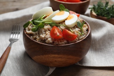 Tasty boiled oatmeal with egg, tomatoes and microgreens served on wooden table, closeup