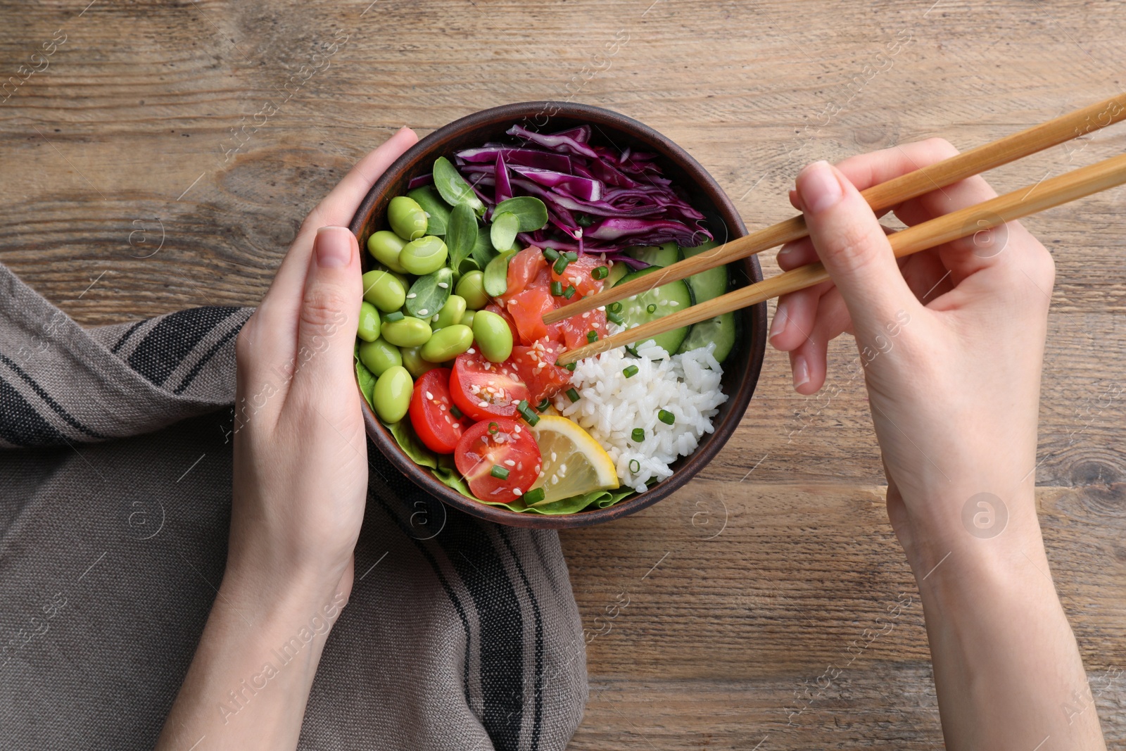Photo of Woman eating poke bowl with salmon, edamame beans and vegetables at wooden table, top view