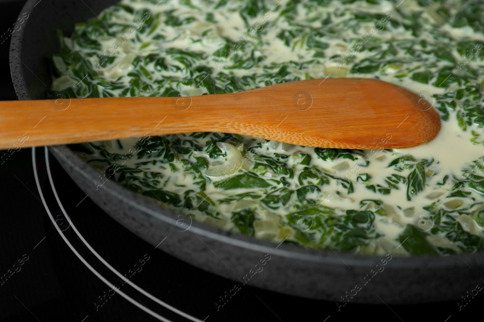 Photo of Tasty spinach dip with wooden spoon in frying pan on kitchen stove, closeup view