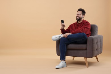 Happy young man using smartphone on armchair against beige background, space for text