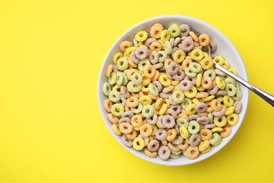 Photo of Tasty cereal rings in bowl and spoon on yellow table, top view. Space for text