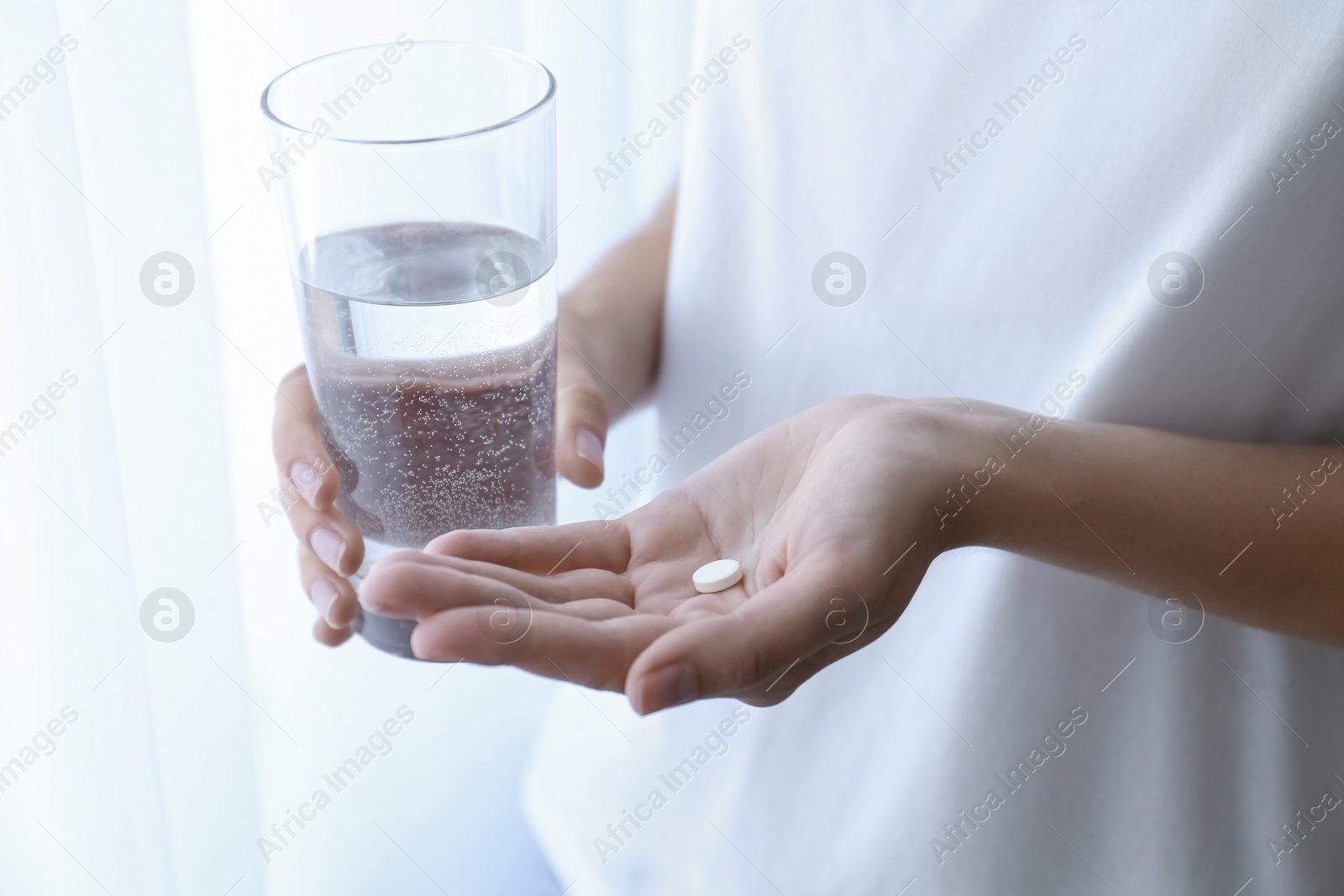 Photo of Woman holding glass of water and pill near window at home, closeup
