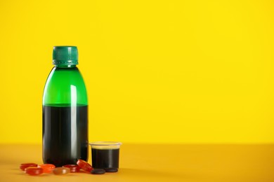 Photo of Bottle of syrup, measuring cup and cough drops on yellow background. Space for text