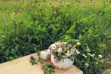 Ceramic mortar with pestle, different wildflowers and herbs on wooden board in meadow