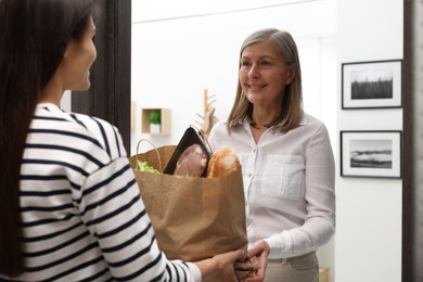 Courier giving paper bag with food products to senior woman indoors