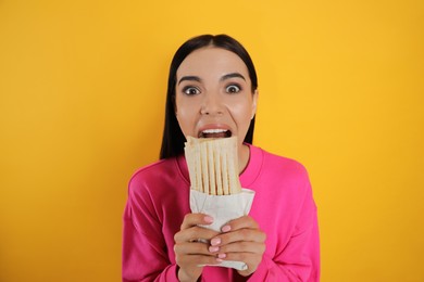 Emotional young woman eating delicious shawarma on yellow background