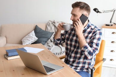Photo of Young man talking on mobile phone while working with laptop at desk. Home office