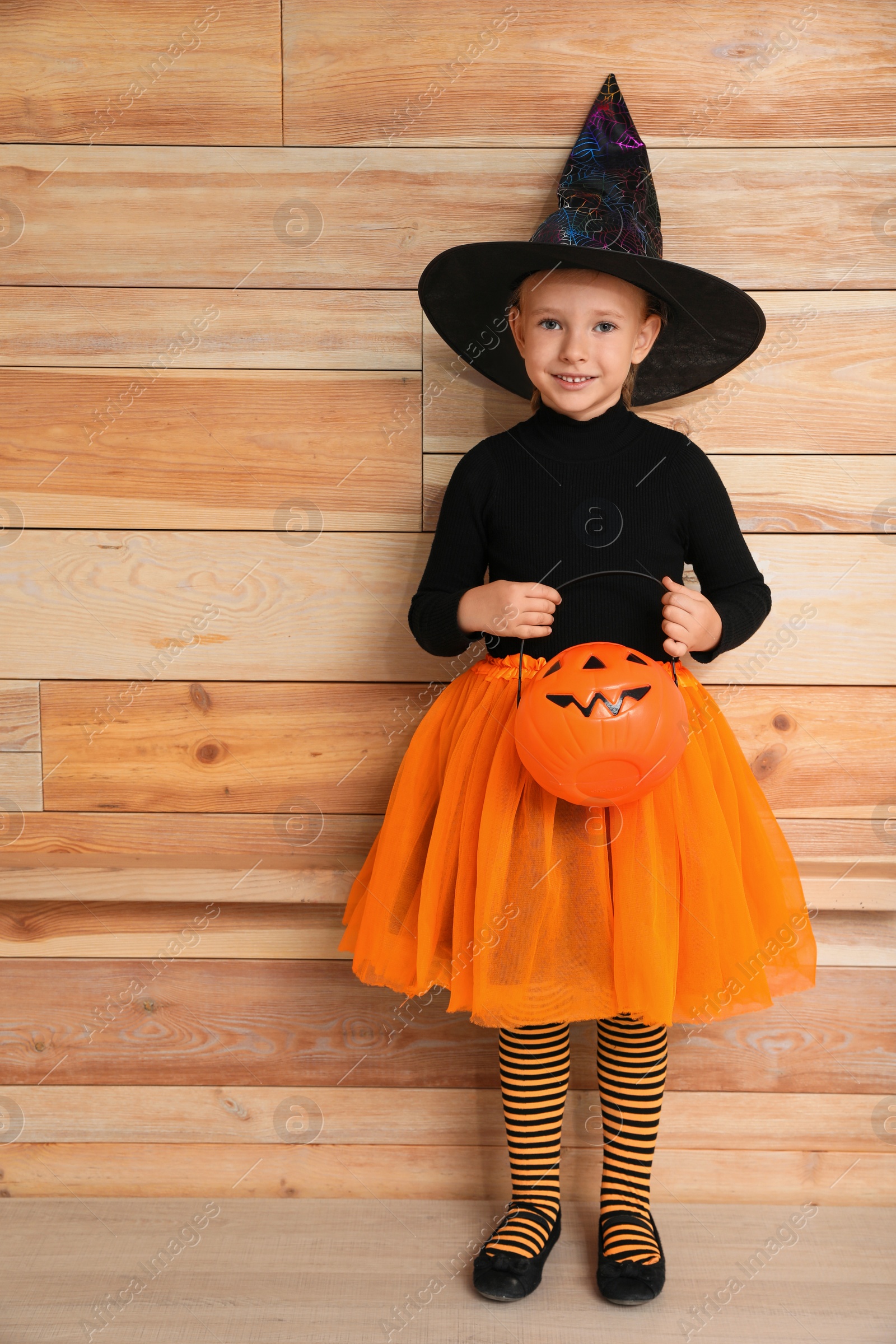 Photo of Cute little girl with pumpkin wearing Halloween costume near wooden wall