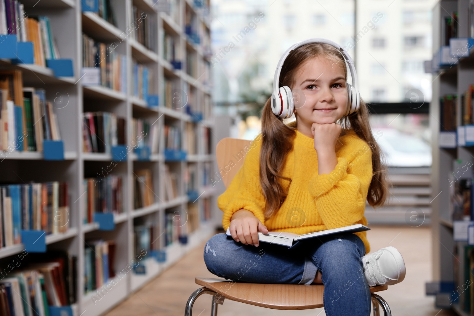 Photo of Cute little girl with headphones reading book on chair in library