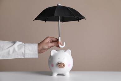 Photo of Woman holding small umbrella over piggy bank against beige background, closeup