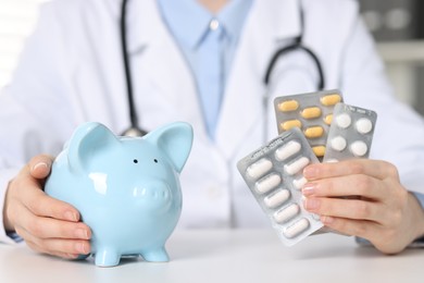 Doctor with pills and piggy bank at white table indoors, closeup