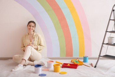 Young woman and decorator's tools near wall with painted rainbow indoors