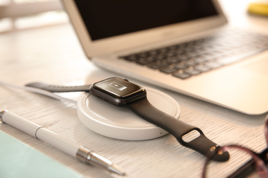 Photo of Smartwatch with wireless charger on white wooden table. Modern workplace accessory