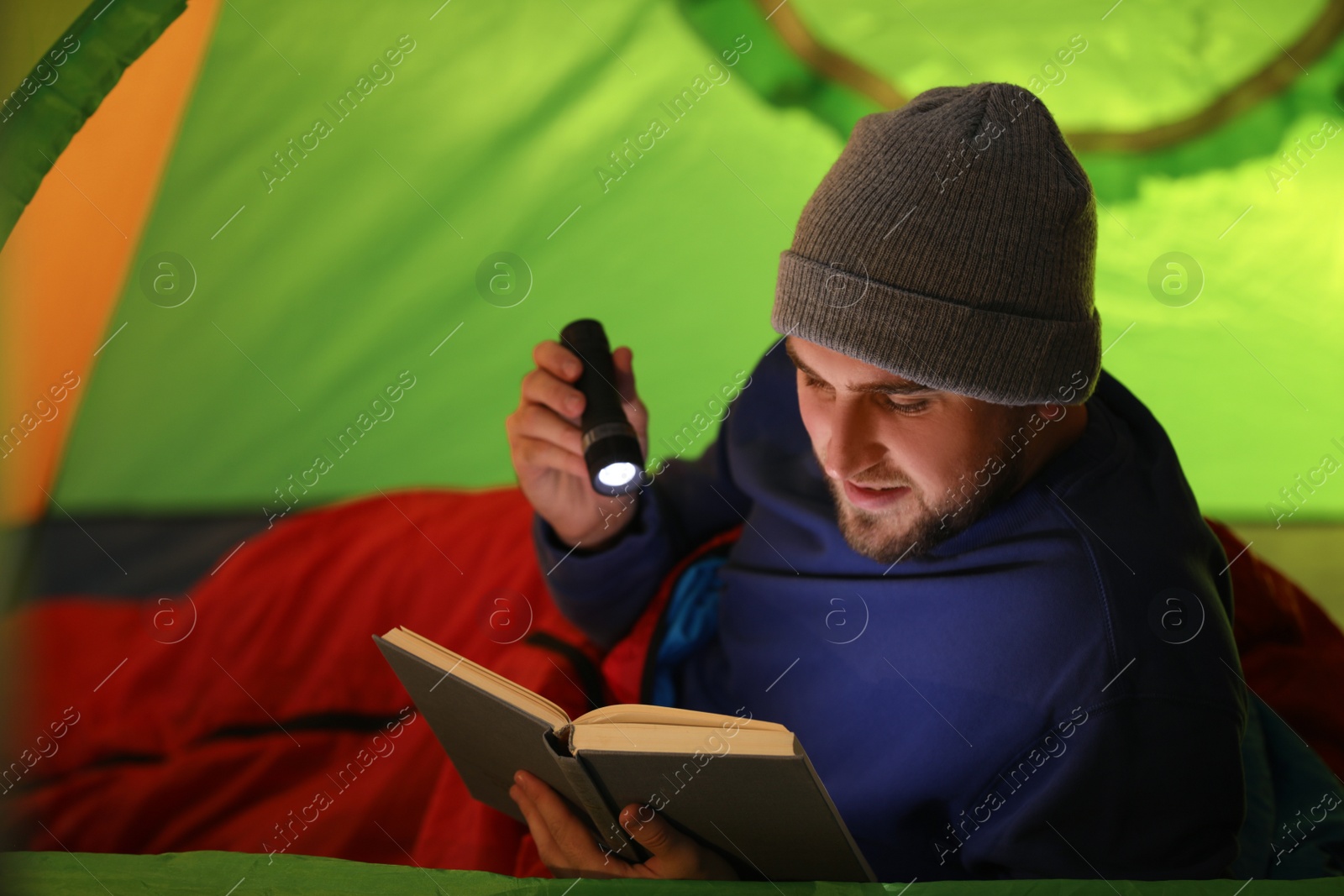 Photo of Young man with flashlight reading book in tent