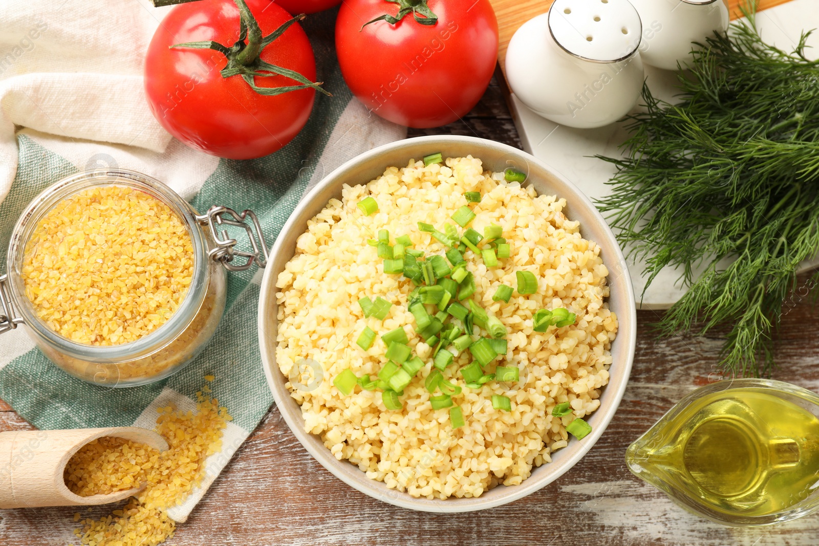 Photo of Delicious bulgur with green onion in bowl, tomatoes, dill and oil on wooden table, flat lay