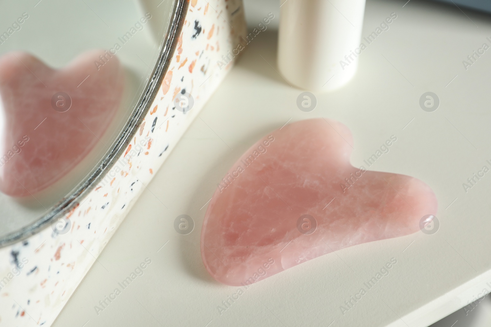 Photo of Rose quartz gua sha tool near mirror on white table, closeup