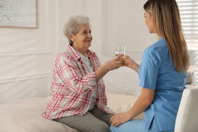 Photo of Young caregiver giving water to senior woman in room. Home care service