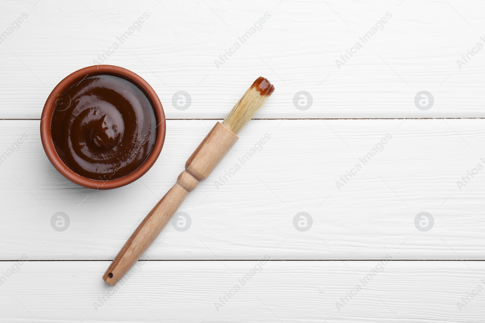 Photo of Tasty barbeque sauce in bowl and brush on white wooden table, top view. Space for text