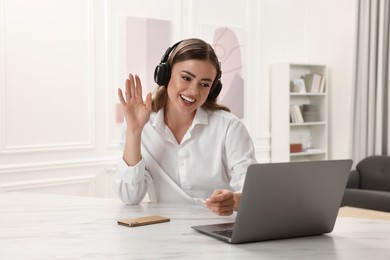 Photo of Happy woman with headphones having video chat via laptop at white table in room