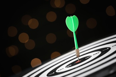 Photo of Green arrow hitting target on dart board against blurred lights. Space for text