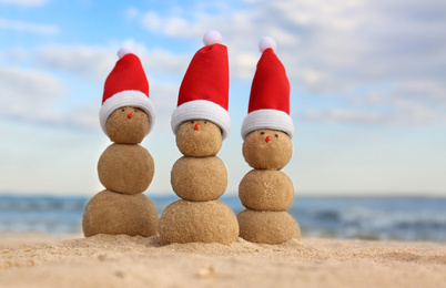 Snowmen made of sand with Santa hats on beach near sea. Christmas vacation