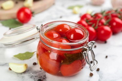 Photo of Glass jar of pickled cherry tomatoes and ingredients on white marble table, closeup