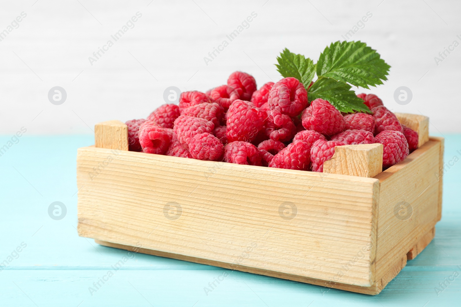 Photo of Crate of delicious fresh ripe raspberries with leaves on blue wooden table