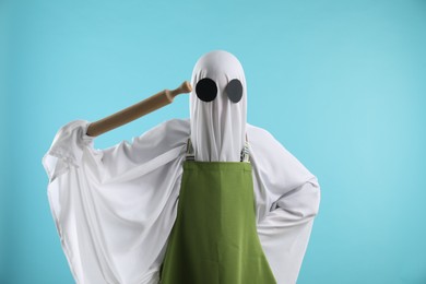 Woman in ghost costume and apron with rolling pin on light blue background