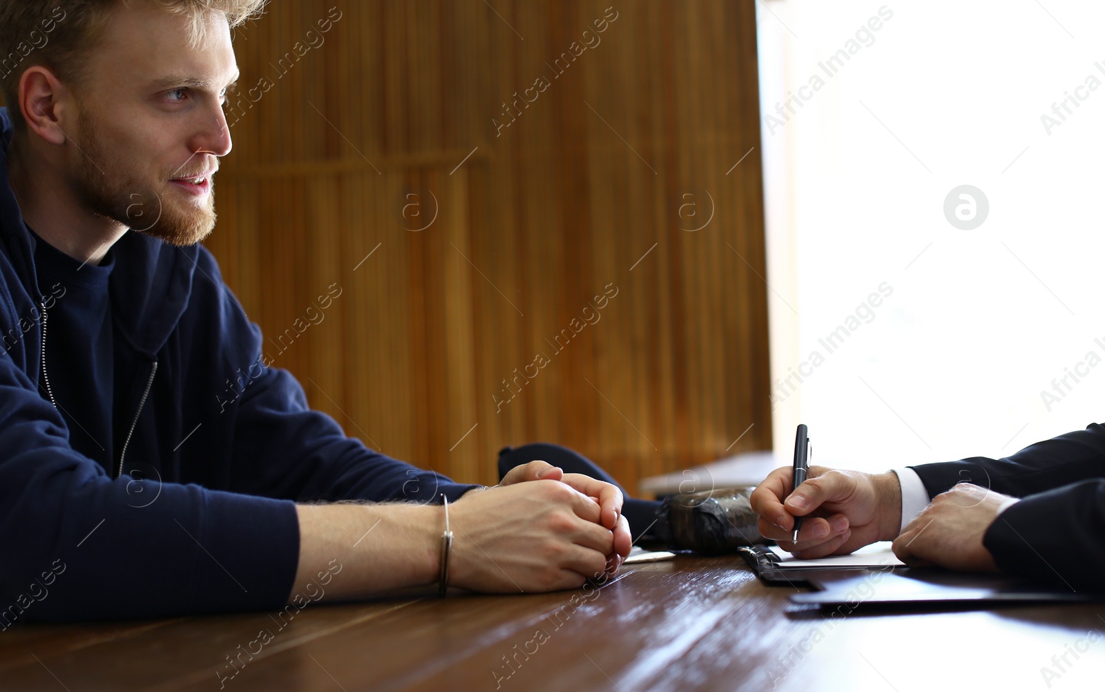 Photo of Police officer interrogating criminal in handcuffs at desk indoors