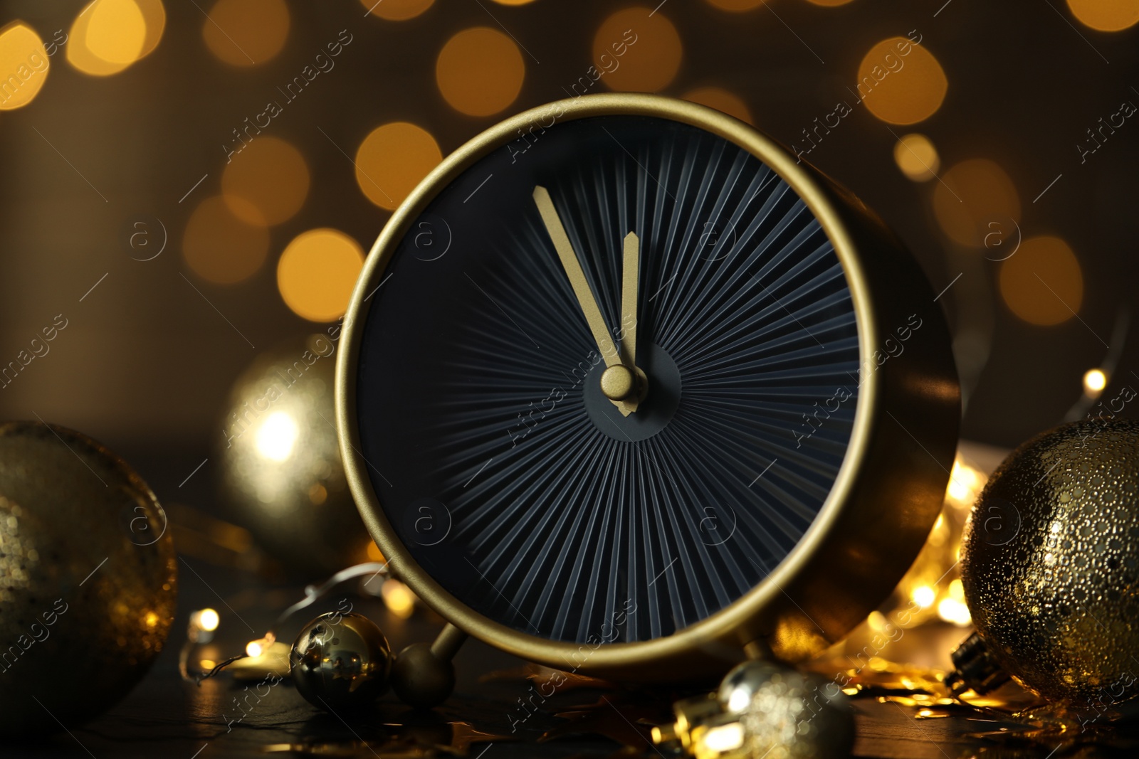 Photo of Stylish clock with decor on black table against blurred Christmas lights, closeup. New Year countdown