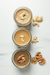 Tasty nut butters in jars and raw nuts on white marble table, flat lay