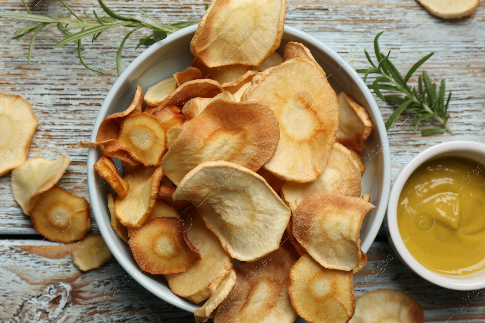 Photo of Tasty homemade parsnip chips with sauce and rosemary on old light blue wooden table, flat lay