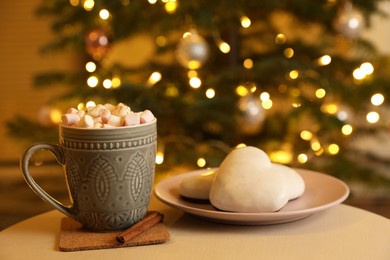 Photo of Cup of tasty cocoa with marshmallows and cookies on white table against blurred Christmas tree