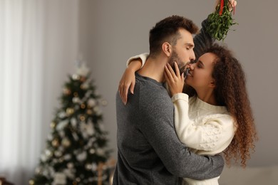 Happy man kissing his girlfriend under mistletoe bunch in room decorated for Christmas