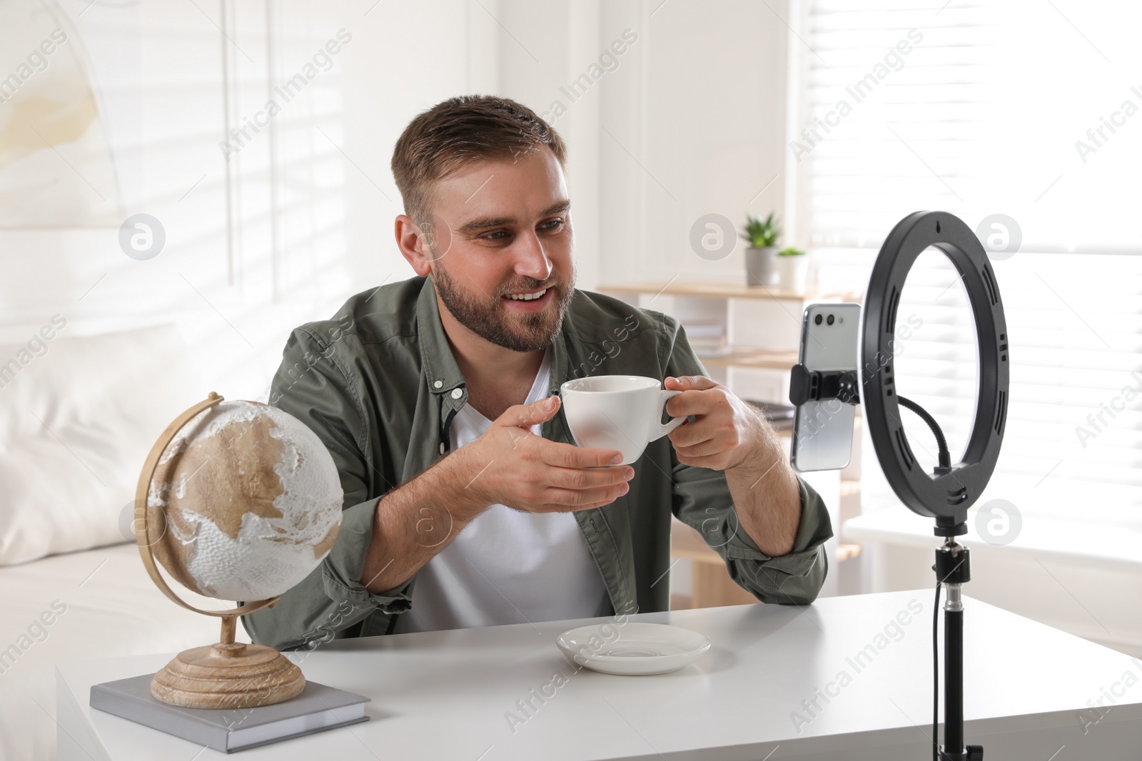 Photo of Blogger with cup of tea recording video at table indoors. Using ring lamp and smartphone