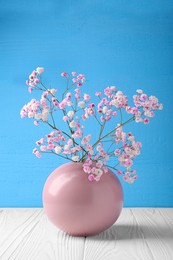 Photo of Beautiful dyed gypsophila flowers in pink vase on white wooden table against light blue background