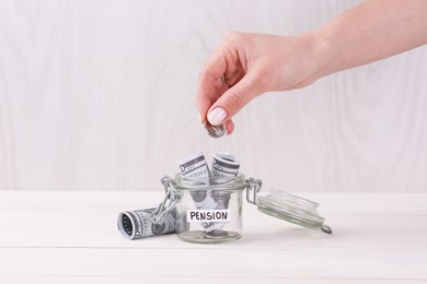 Photo of Pension concept. Woman putting coin into glass jar with money at white wooden table, closeup