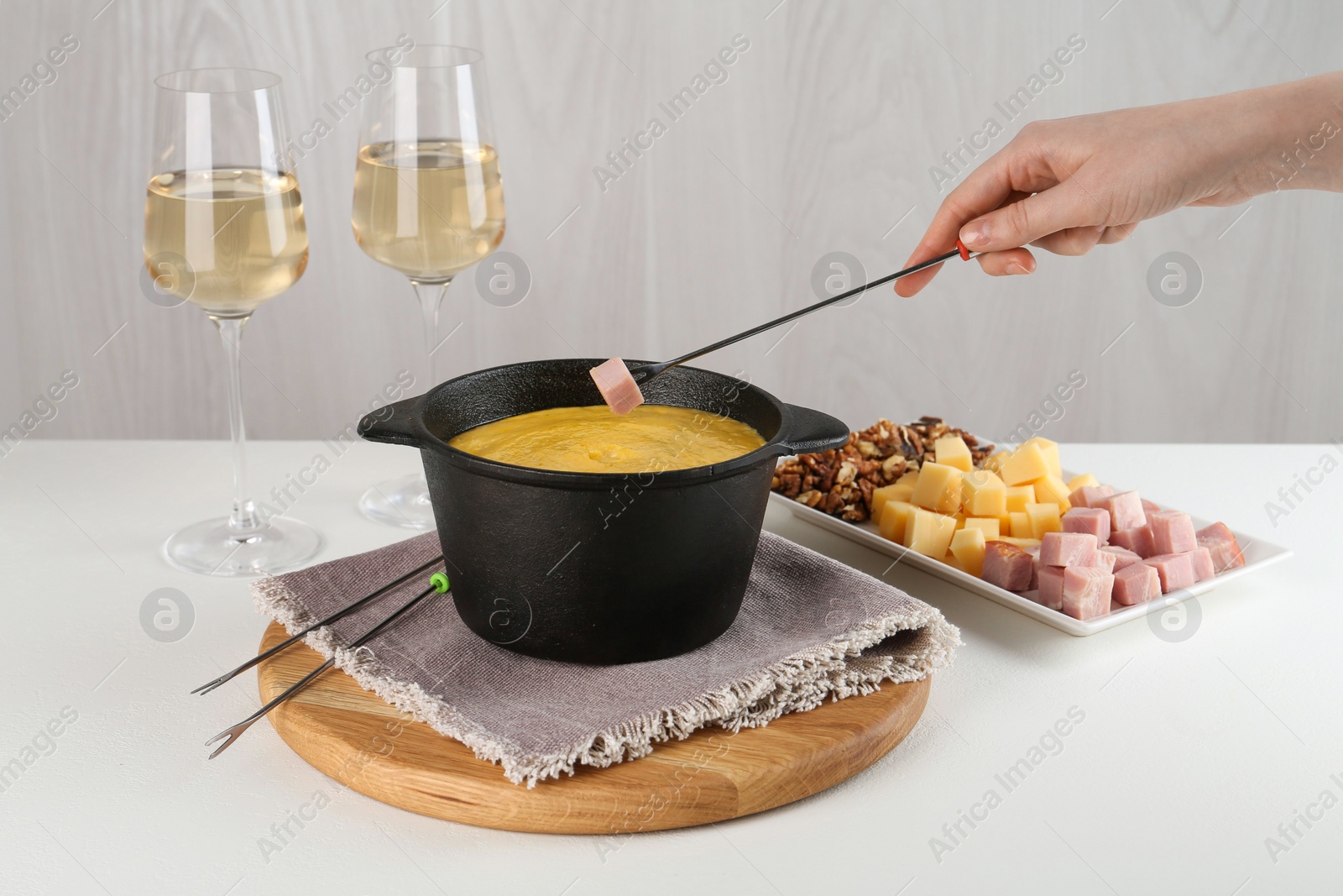 Photo of Dipping piece of ham into fondue pot with tasty melted cheese at white table, closeup