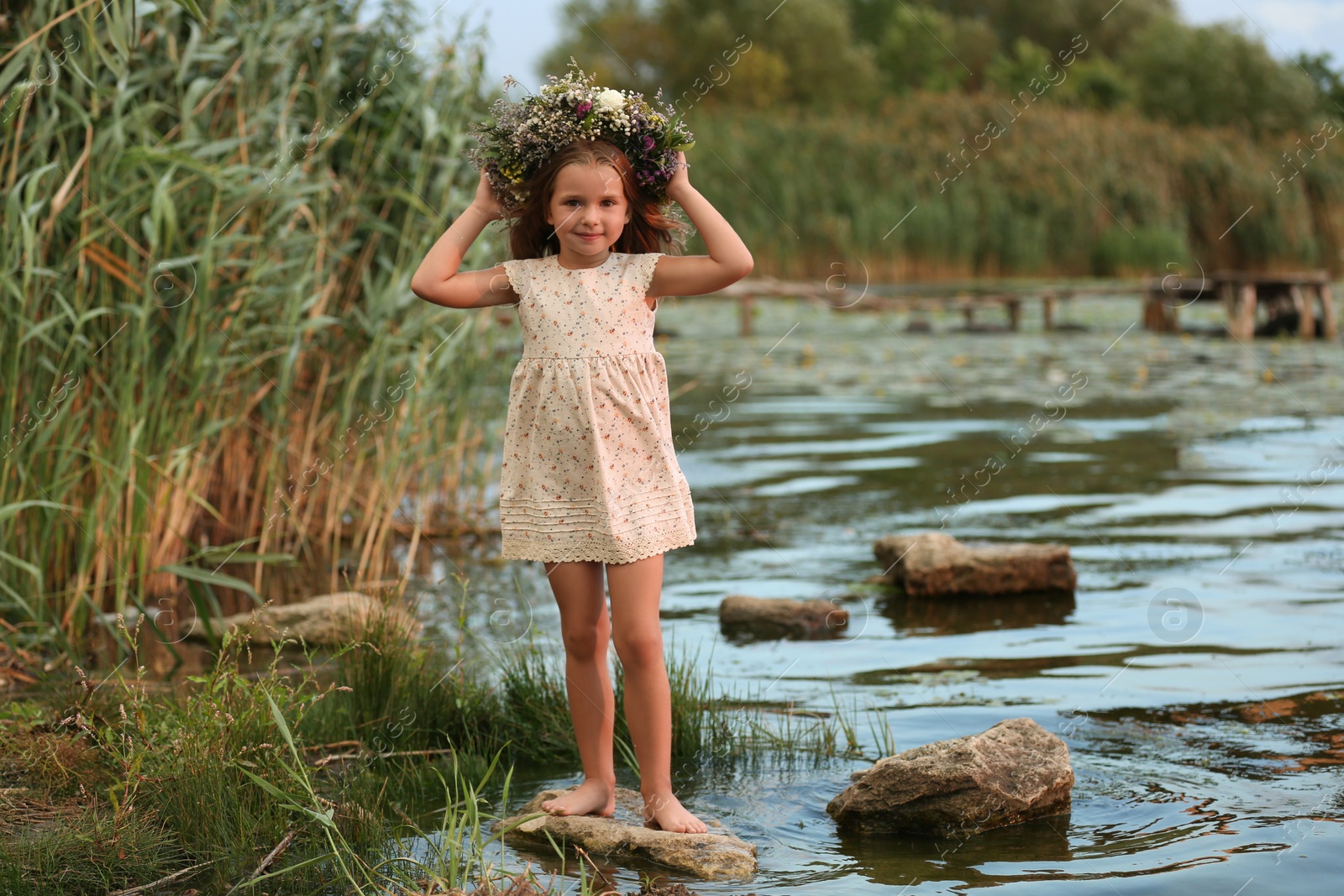 Photo of Cute little girl wearing wreath made of beautiful flowers near river