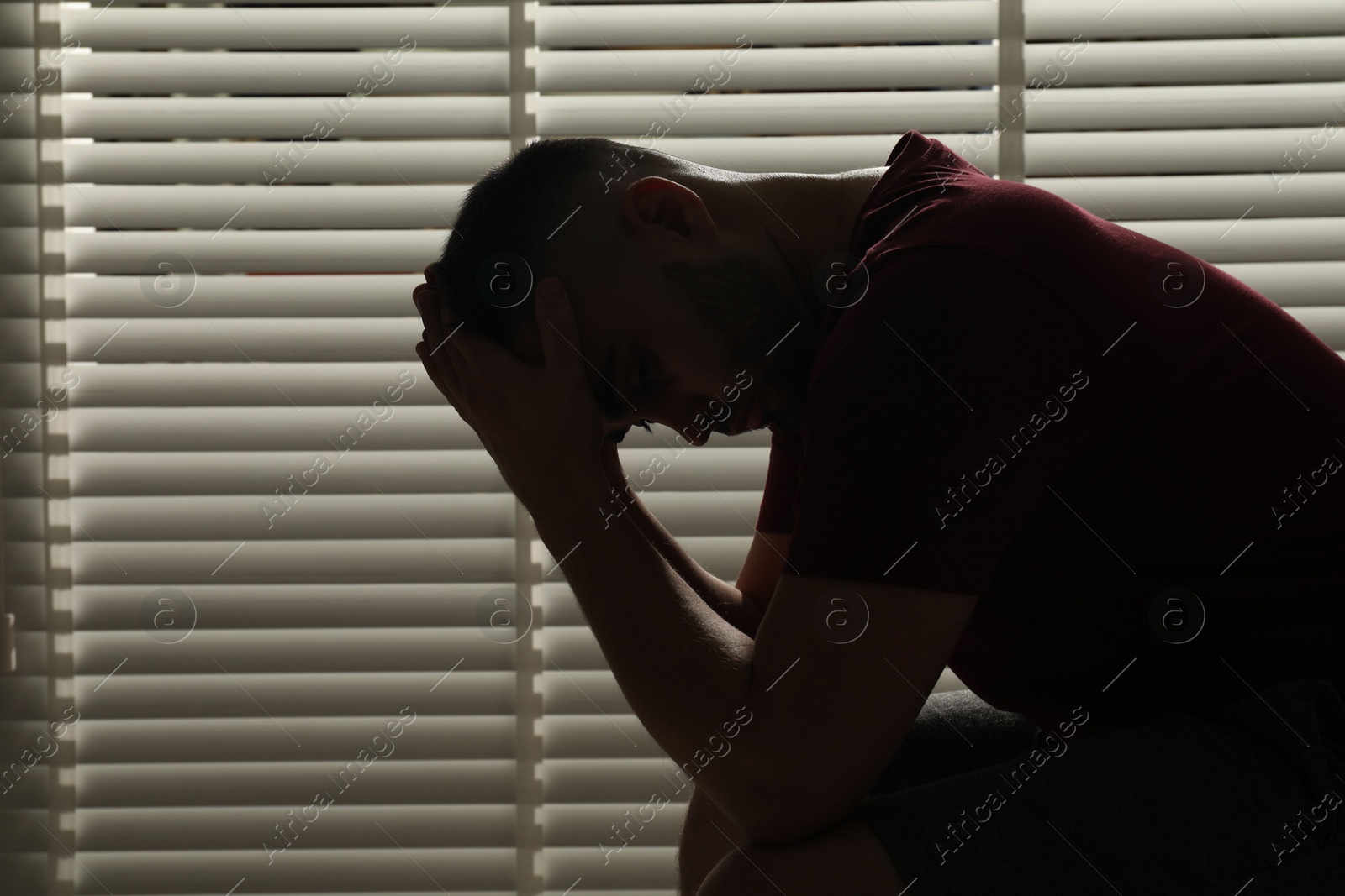 Photo of Sad man sitting near closed blinds indoors. Space for text